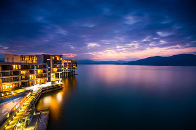 Illuminated hotel by lake against cloudy sky at dusk