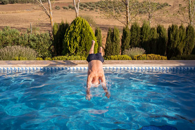 Full body back view of unrecognizable barefoot and shirtless senior male jumping in swimming pool with clear blue water