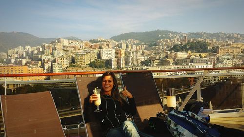 Young woman gesturing thumbs up sign with summer drink while sitting on deck chair against cityscape