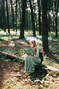 Woman wearing hijab reading book while sitting in forest