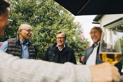 Smiling elderly male friends talking while standing at back yard during garden party