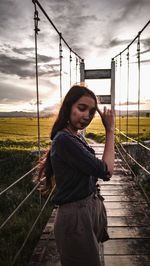 Side view of young woman standing on railing against sky