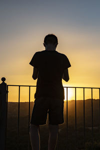 Rear view of silhouette man standing by railing against sky during sunset