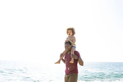 Happy man carrying daughter on shoulders standing in front of sea at beach