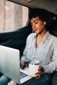 Young content cheerful african american female working with netbook and having mug of coffee to go riding on backseat in car
