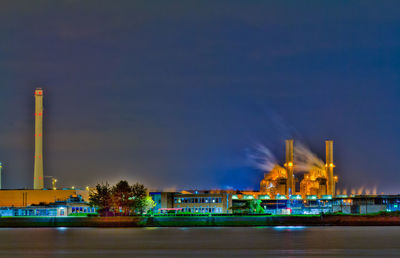 Illuminated factory by river against sky at night