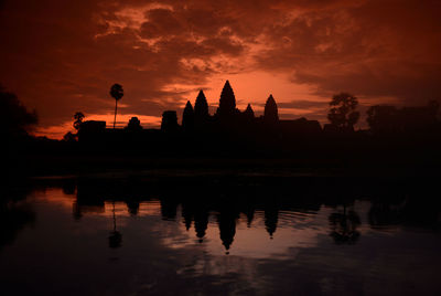 Reflection of silhouette built structure and trees in lake against orange sky during sunset