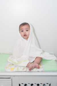 Portrait of cute baby boy wrapped in towel while sitting on bed against wall