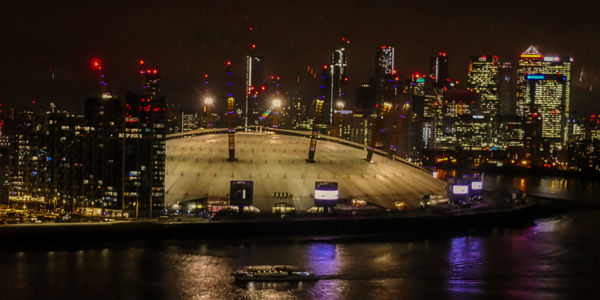 Illuminated modern buildings by river against sky at night