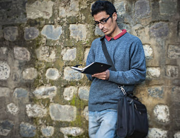 Man reading book while standing by stone wall