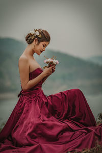 Side view of young woman holding flowers while sitting against sky