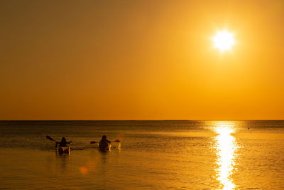 Silhouette friends kayaking in sea against clear orange sky