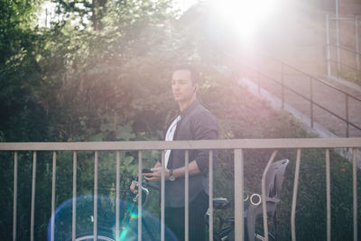 Man standing with mobile phone and bicycle on footbridge against trees