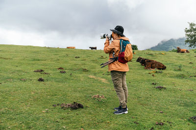 Female photographer taking pictures with a camera standing in the green meadow with grazing cows.