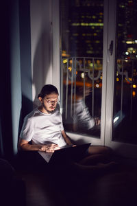 Freelance man in glasses working, sits at the window on the floor in front of a skyscraper at night