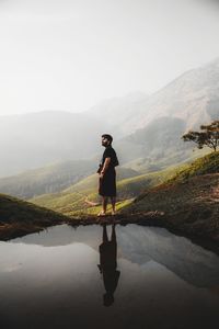 Full length of man standing by lake while looking at mountains during foggy weather