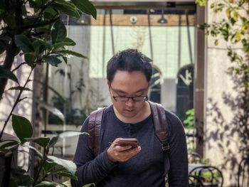 Young man using mobile phone behind plant in courtyard against reflective glass windows.