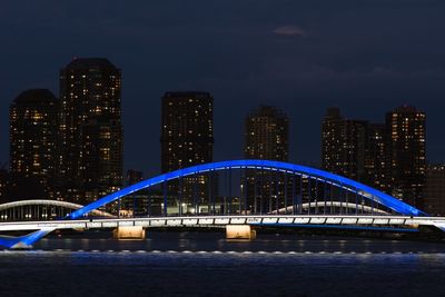 Illuminated bridge over river by buildings against sky at night