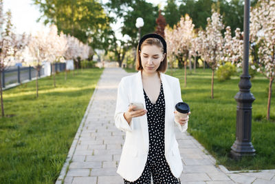 Young woman holding coffee using smart phone while standing on footpath