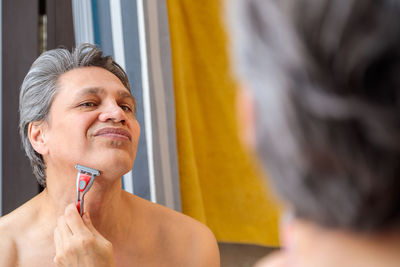 An adult gray-haired man shaves with a razor in front of a mirror