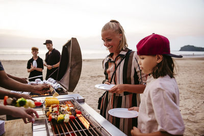 Mother and daughter standing near barbecue grill while holding plates at beach