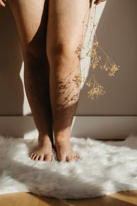 Bare legs of crop anonymous barefoot female with dried twig standing near wall on white rug in light room with sunlight