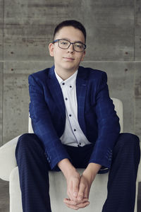 A boy with glasses with an intelligent look sits in a chair with his hands clasped in a lock