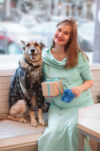 Pregnant woman is sitting in cafe with her dog and holding gift box and blue knitted bootee.