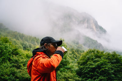 Man in orange jacket drinking from cup against foggy mountains