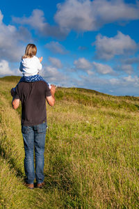 Father carrying daughter on shoulder while walking on grassy field against sky