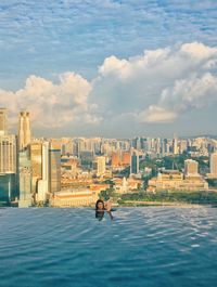 Woman swimming in infinity pool against cityscape and cloudy sky