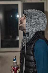 Side view of young man in hood drinking beer indoors