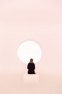 Rear view of a woman sitting in front of a light pastel colored circle on the wall.