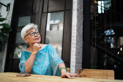 Thoughtful senior woman with mobile phone on table looking away while sitting at sidewalk cafe