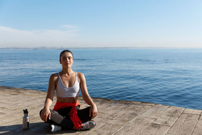 Full length of young woman sitting at sea shore against sky