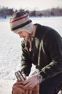 Young man putting thermos in bag while kneeling on snow covered field