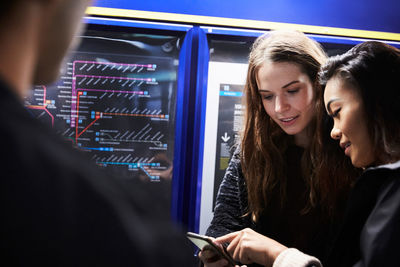 Female friends looking at smart phone while standing by illuminated map in subway station