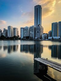 Modern buildings by river against sky during sunset