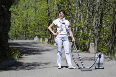 Full length of woman with vacuum cleaner standing on road amidst trees