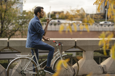 Young man sitting on bicycle and sending voicemail by railing