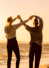 Rear view of two friends holding hands while standing at beach sunset