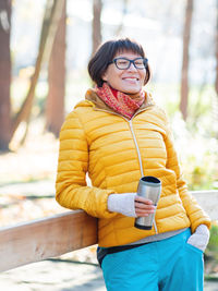 Wide smiling women in yellow jacket is holding thermos mug. hot tea or other beverage on cool day.