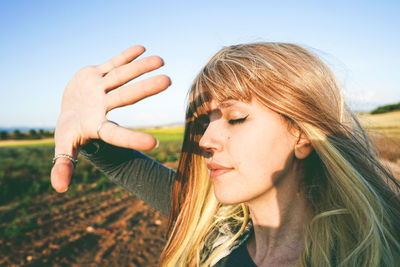 Young woman shielding eyes while standing at field against sky on sunny day