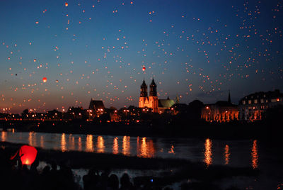 Sky lanterns flying over river by city during dusk