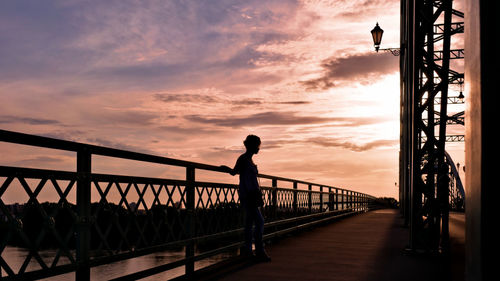 Silhouette woman standing on footbridge against sky during sunset