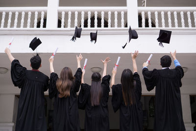 Rear view of students in university gowns throwing mortarboards while standing against building