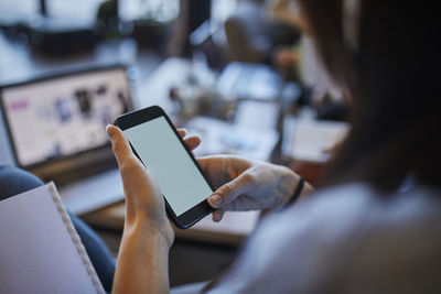 Young woman in cafe using smartphone with laptop in background