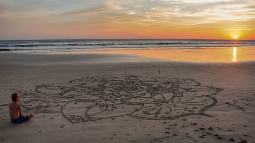 Shirtless man sitting by drawing at beach against sky during sunset