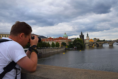 Photographer photographing bridge while leaning on railing against cloudy sky