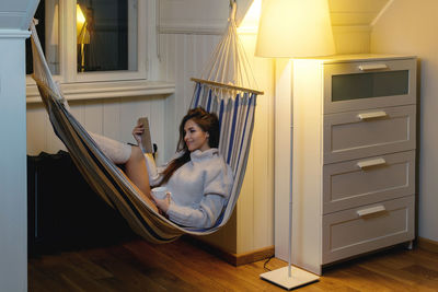 Smiling woman using phone sitting in hammock at home